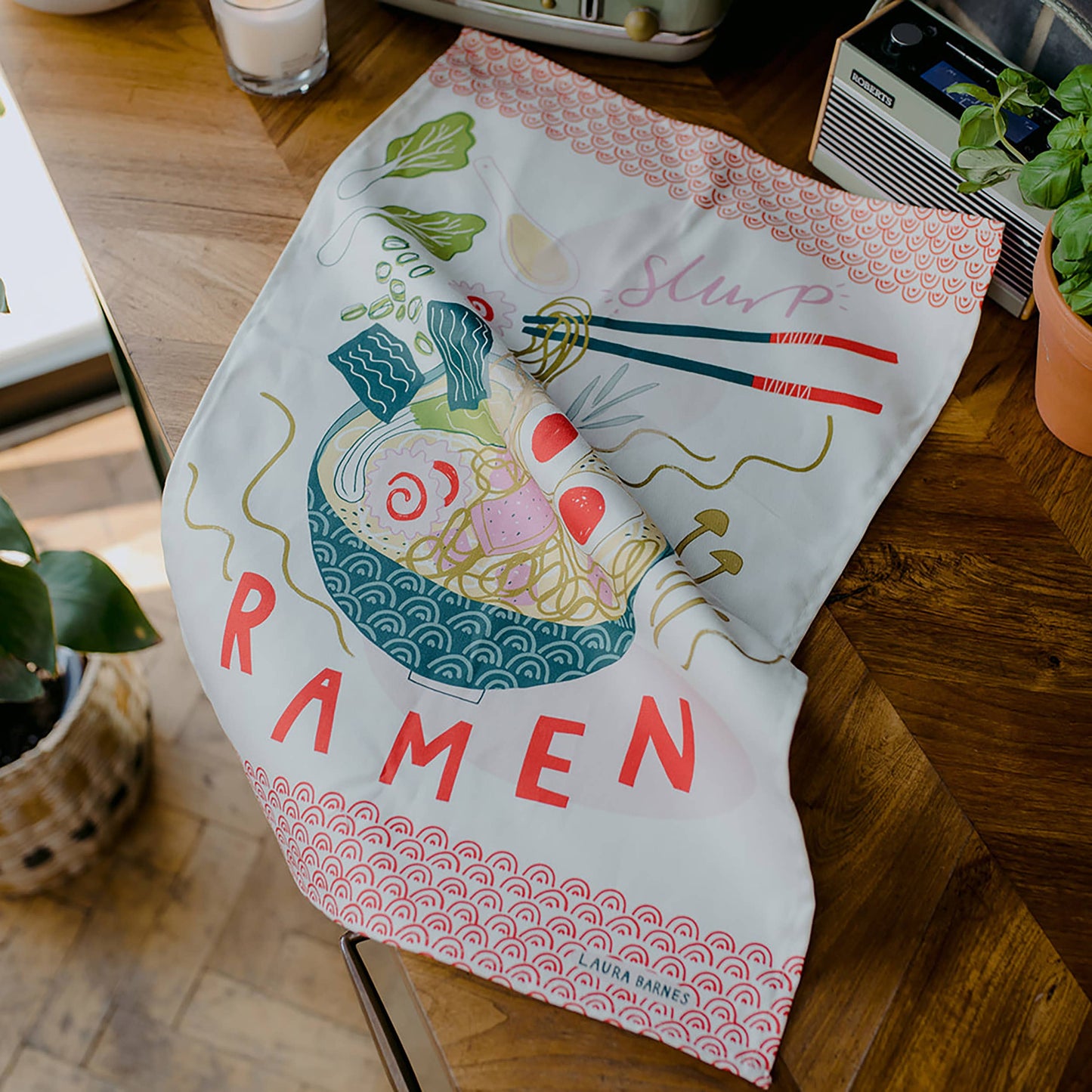 A tea towel illustrated with ramen and chopsticks sitting on a wooden kitchen table.