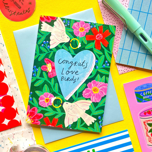 Colorful stationery items including a 'congrats love birds' card with birds and flowers, a pen, and various stationery pieces on a bright background.