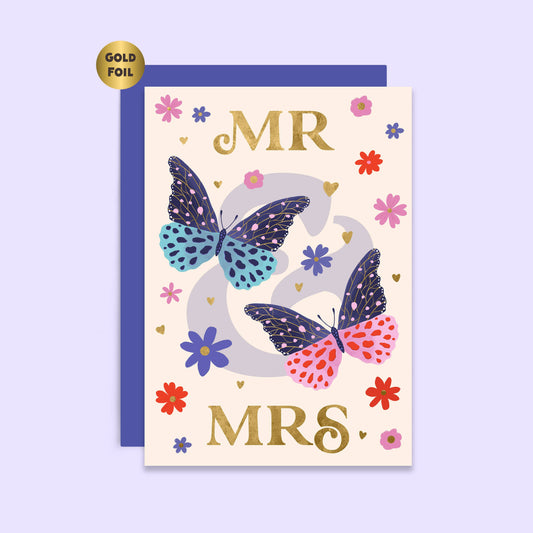 A wedding card with 'MR & MRS' text in gold foil, accompanied by gold foil butterflies and colorful flowers on a cream background. A blue envelope is visible behind the card.