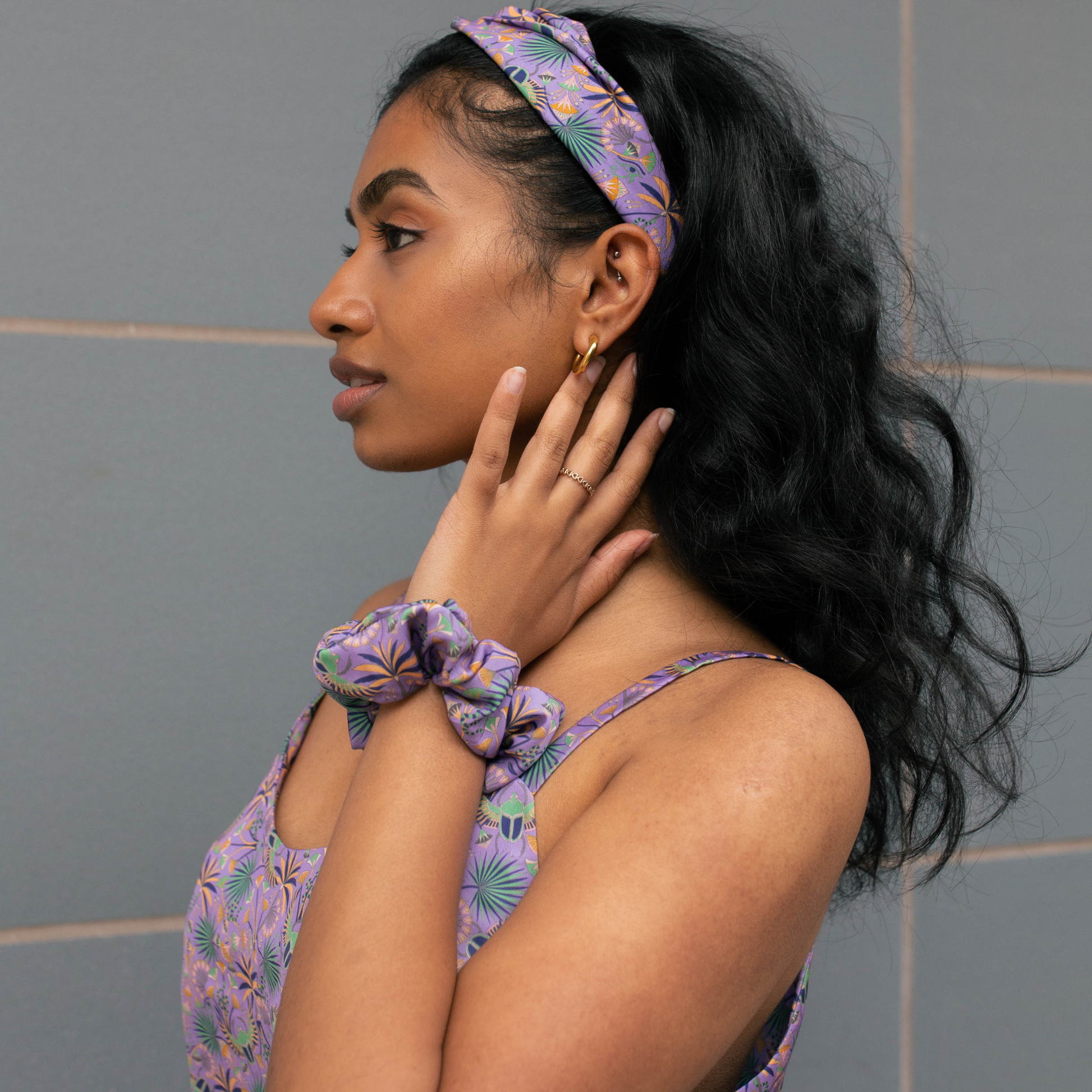 Woman wearing a purple floral headband and scrunchie against a gray tiled wall.