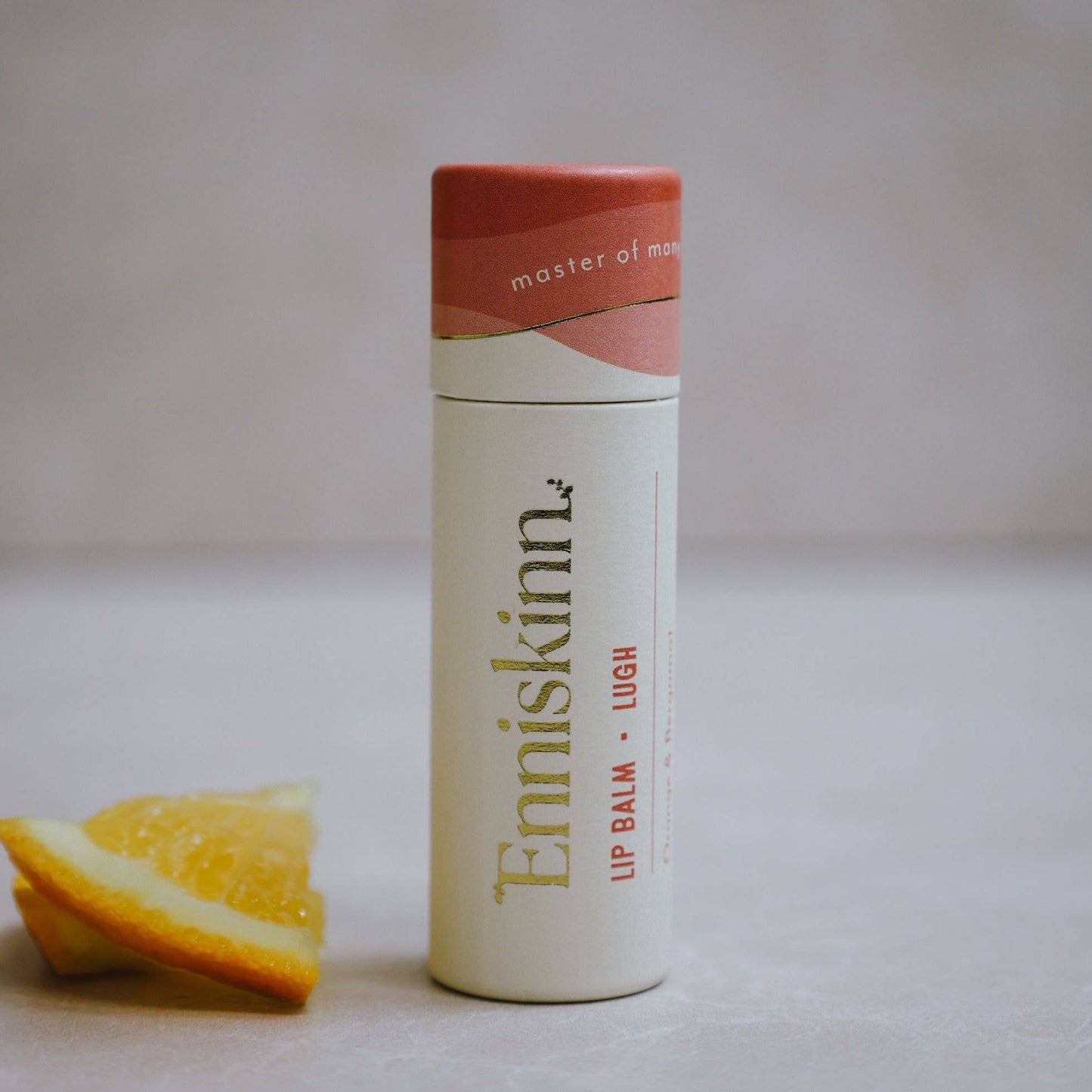 A stick of 'Enniskinn - Lugh' lip balm with orange and bergamot, placed upright beside a slice of orange, with the brand name and product scent visible on the packaging.