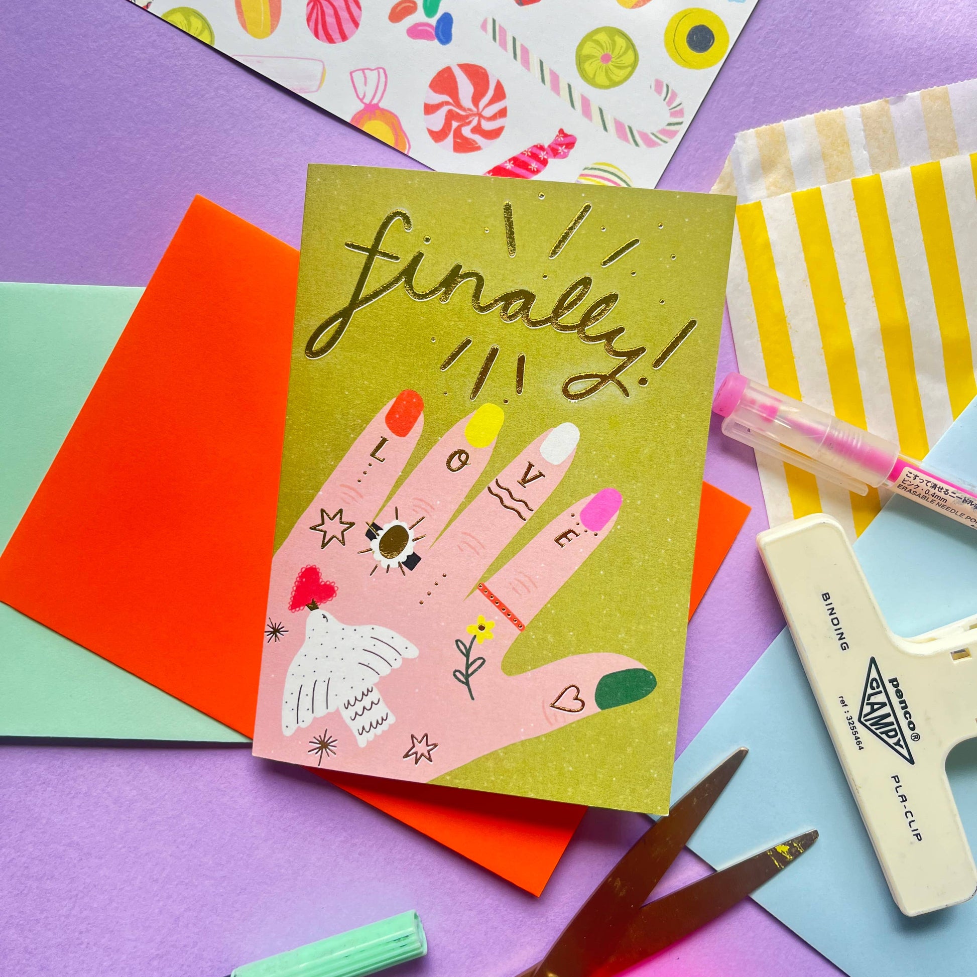 A greeting card with a colorful hand-drawn design of a tattooed hand wearing an engagement ring, featuring the word 'Finally!' in a celebratory tone, placed on a table alongside various colorful crafting materials.