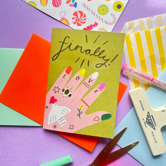 A greeting card with a colorful hand-drawn design of a tattooed hand wearing an engagement ring, featuring the word 'Finally!' in a celebratory tone, placed on a table alongside various colorful crafting materials.