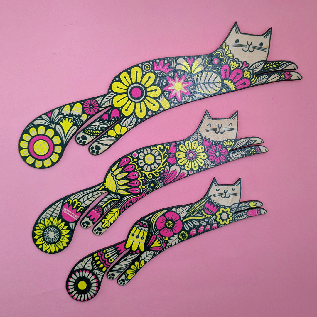 A set of three colorful wooden cutouts of cats with floral patterns on a pink background.