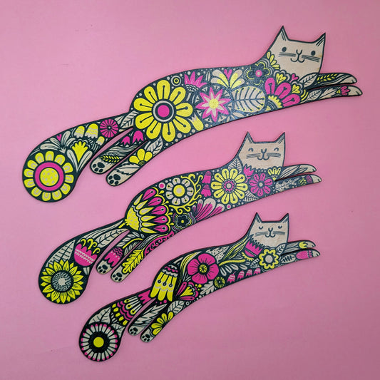 A set of three colorful wooden cutouts of cats with floral patterns on a pink background.