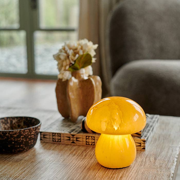 Yellow mushroom-shaped lamp on a wooden surface with a vase and flowers in the background.