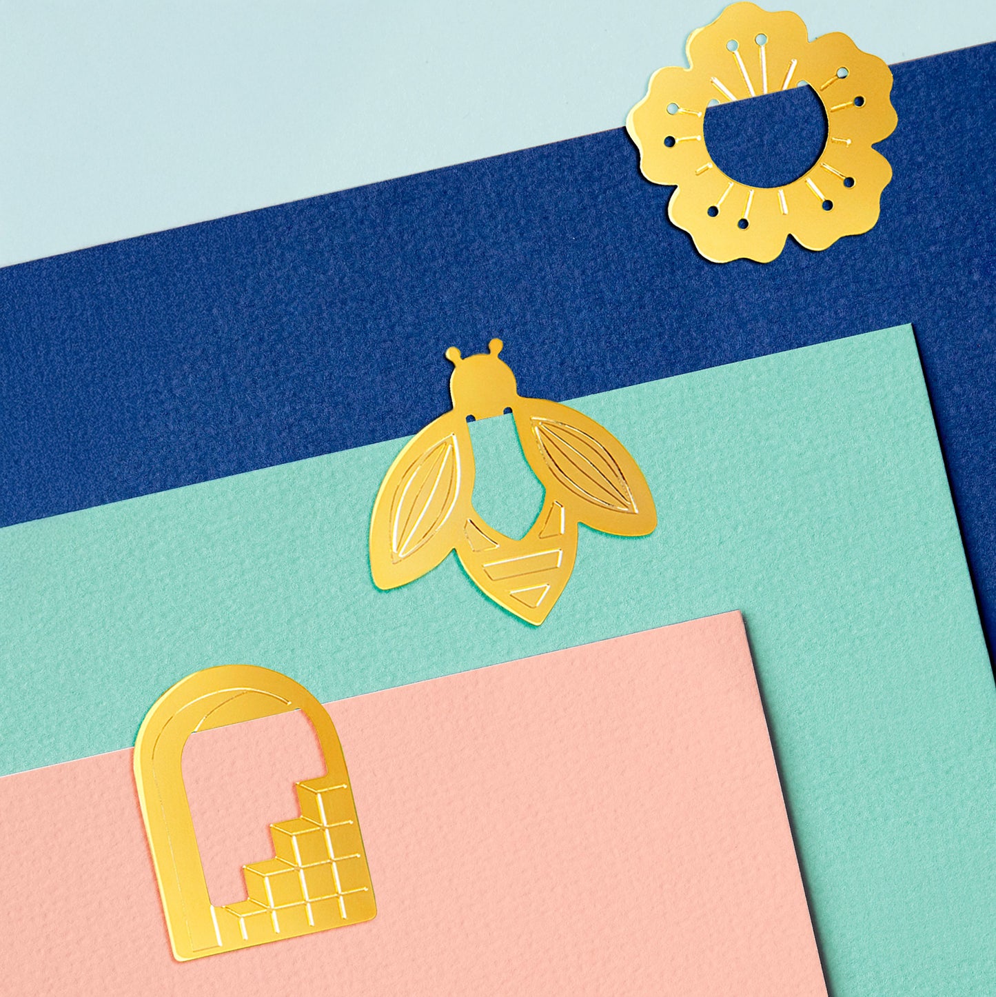 brass coloured paper clips in the shape of a doorway, a bee and a flower, clipped on to bright coloured card.