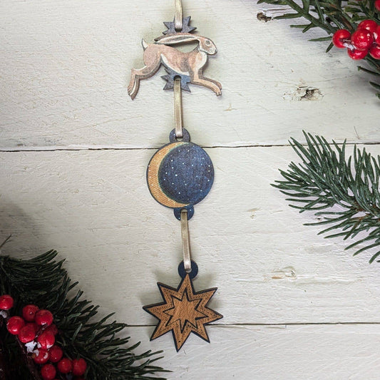 Decorative hanging ornament with hare, moon, and star on a wooden surface with greenery and berries.