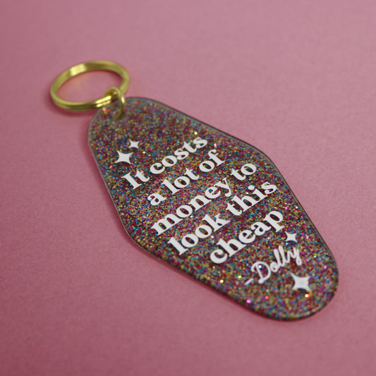 Glittery keychain with text reading "IT COSTS A LOT OF MONEY TO LOOK THIS CHEAP" in a pink background