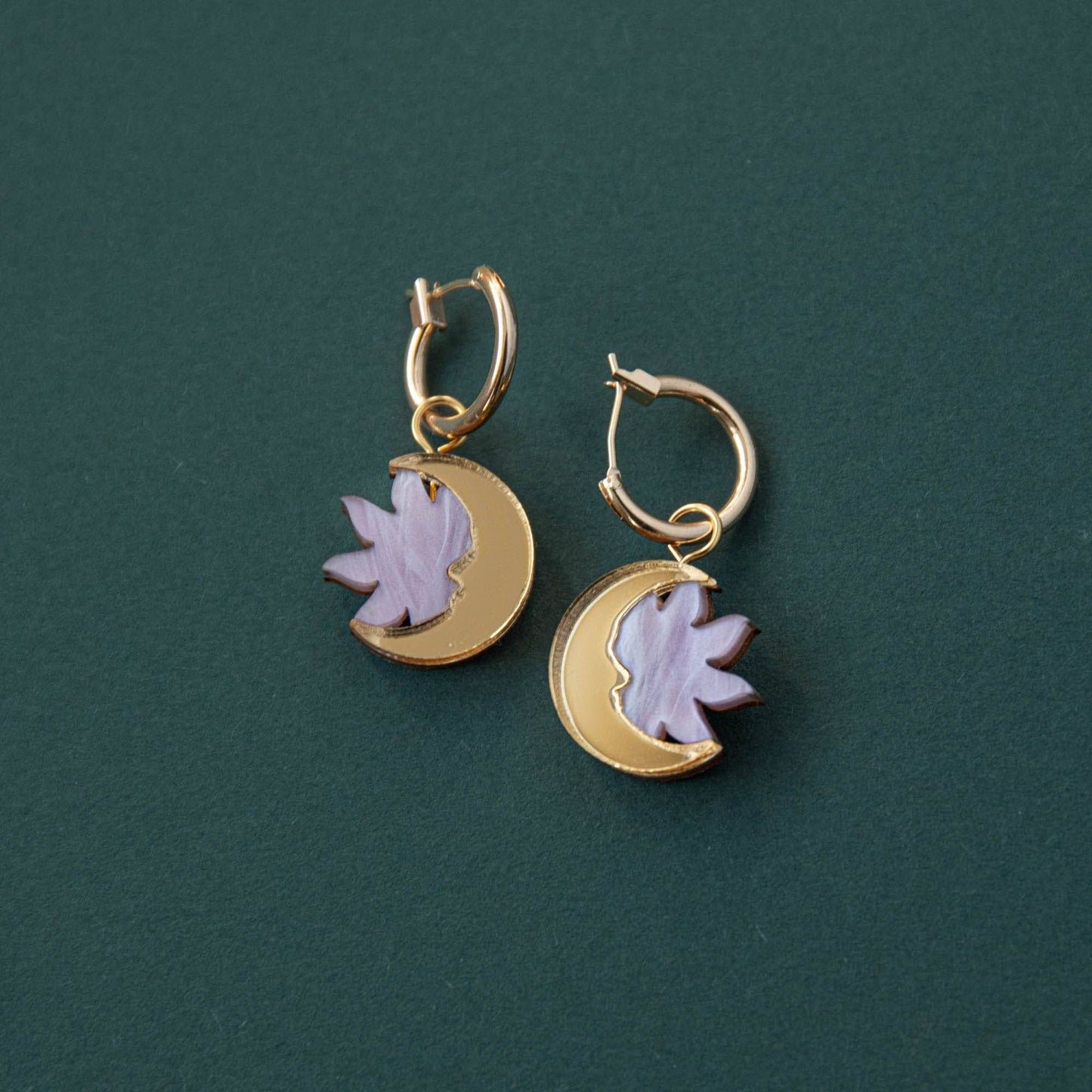A pair of lilac marble and gold hoop earrings with a sun and moon design, displayed against a green background.