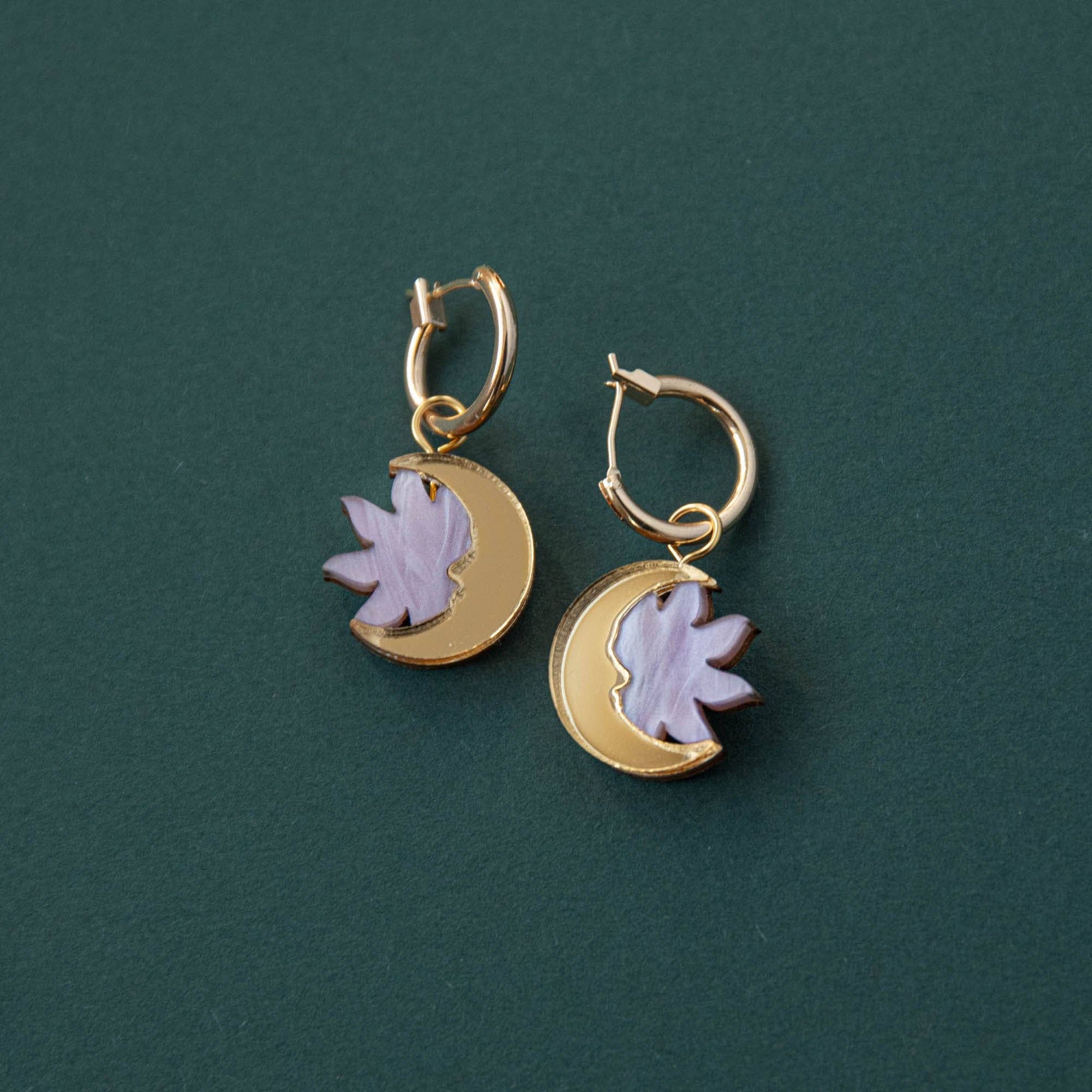 A pair of lilac marble and gold hoop earrings with a sun and moon design, displayed against a green background.