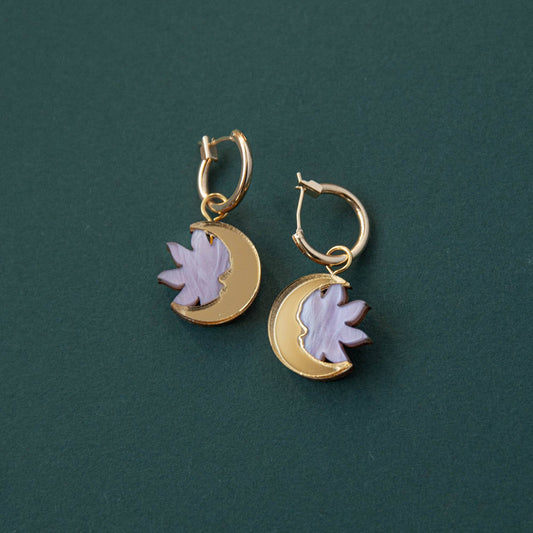 A pair of lilac marble and gold hoop earrings with a sun and moon design, displayed against a green background.
