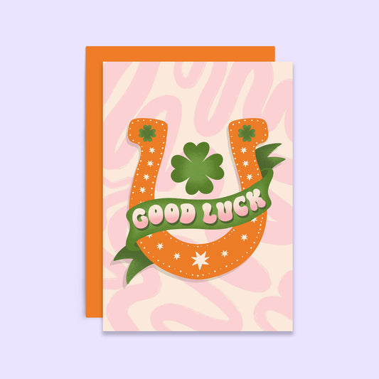 A greetings card with a horseshoe and clovers design on a pink background, accompanied by an orange envelope. The card has a blank inside and is decorated with a retro style.