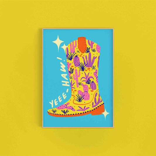 A colorful art print of a cowboy boot with pink cacti on a blue background, with the text 'Yeehaw!' at the top. Print sits on a bright yellow background.