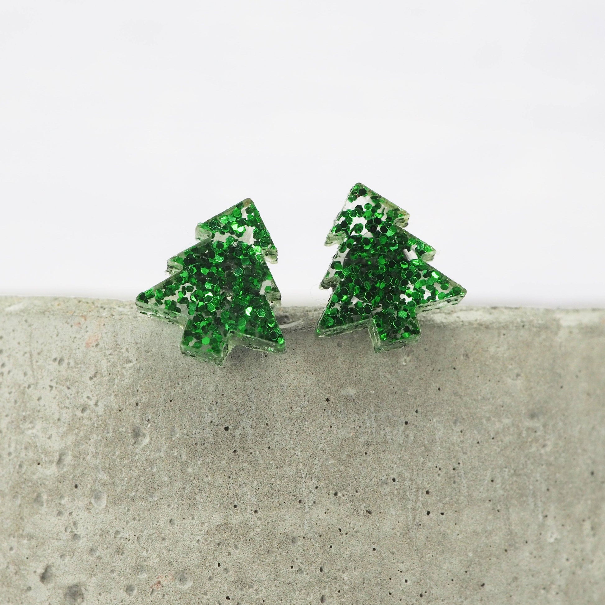 Green glittery Christmas tree earrings on a concrete surface with a white background