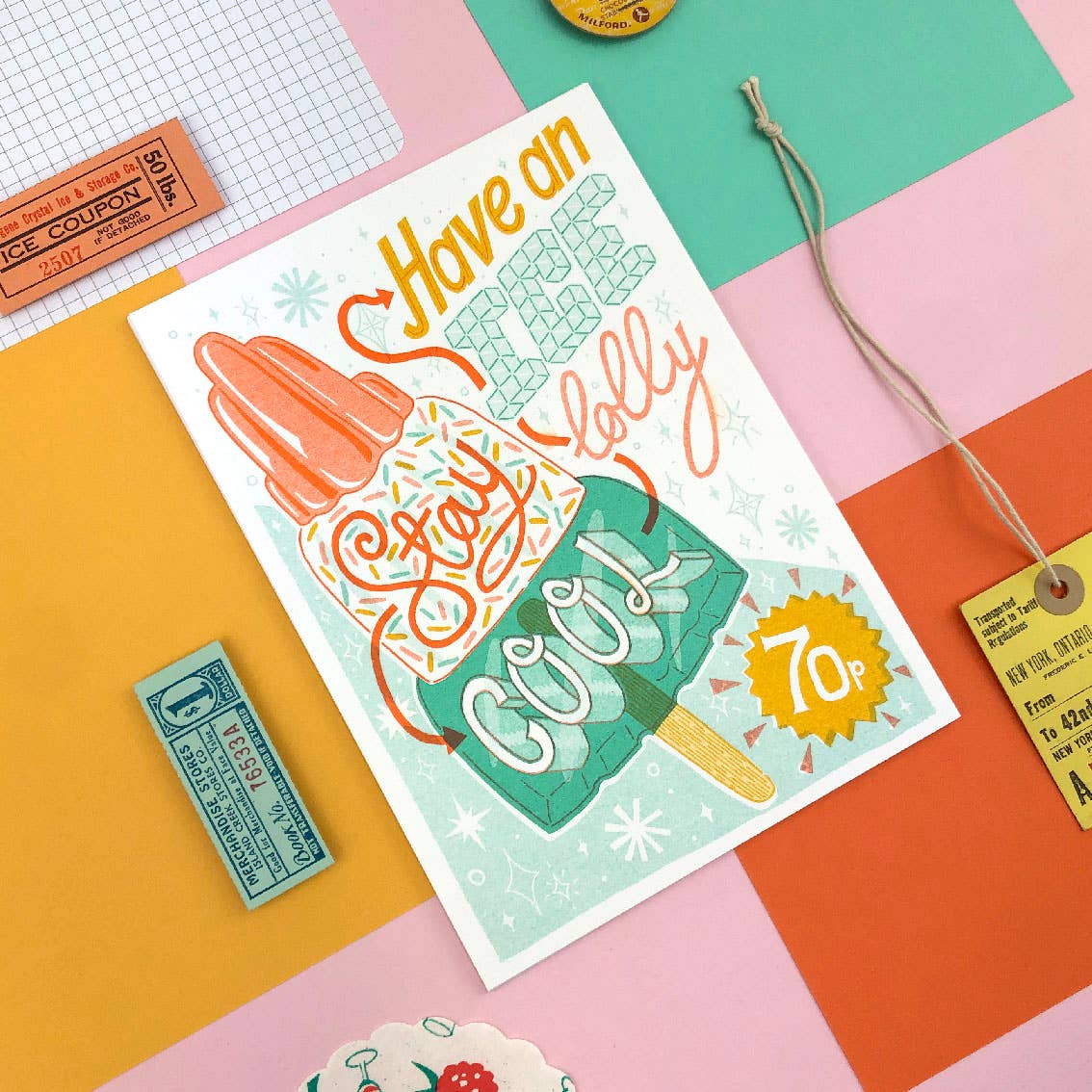 An A5 sized art print laid flat, featuring an illustration of a lollipop with text around it, on a colorful background. There are also small, vintage-style price tags and a string laid out next to the print.