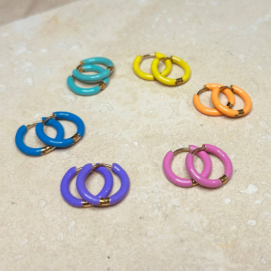 A collection of colorful enamel hoop earrings in blue, green, yellow, orange, pink, and purple, displayed on a sandy surface.