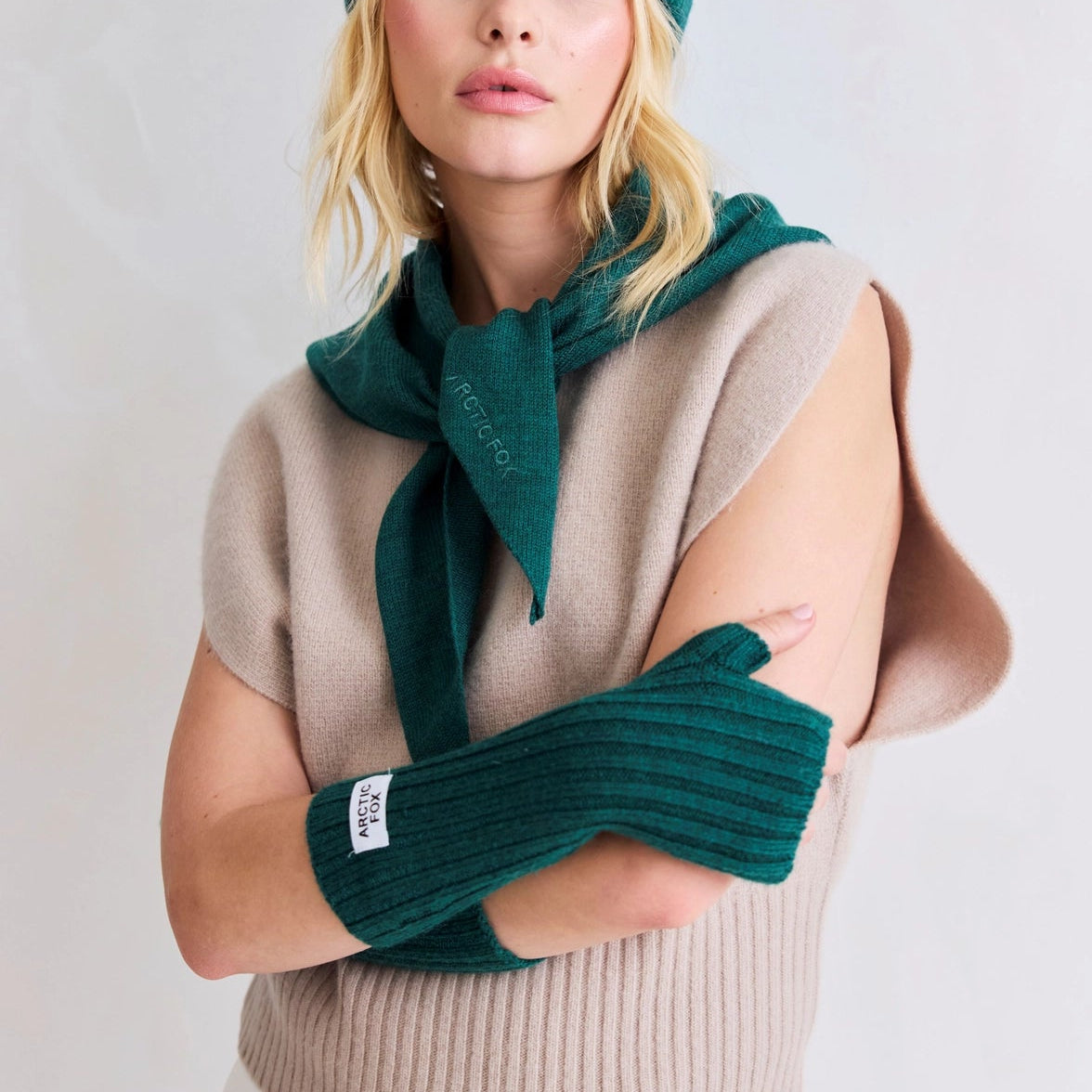 Woman wearing a green knit hat, scarf, and gloves with a beige sweater on a white background