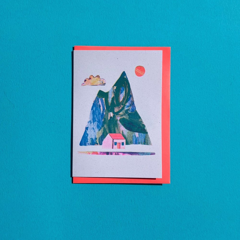 A greeting card with a colorful mountain and house design, accompanied by a red envelope, displayed against a blue background.