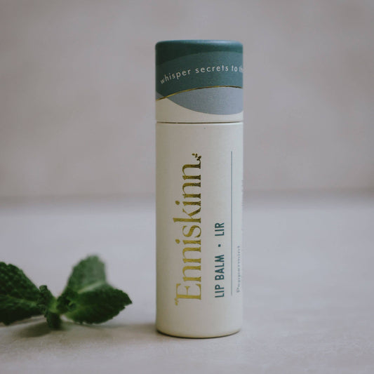 A stick of Enniskinn lip balm with peppermint scent, displayed next to a sprig of peppermint. The packaging is predominantly white with green accents and text, indicating the natural peppermint flavor.
