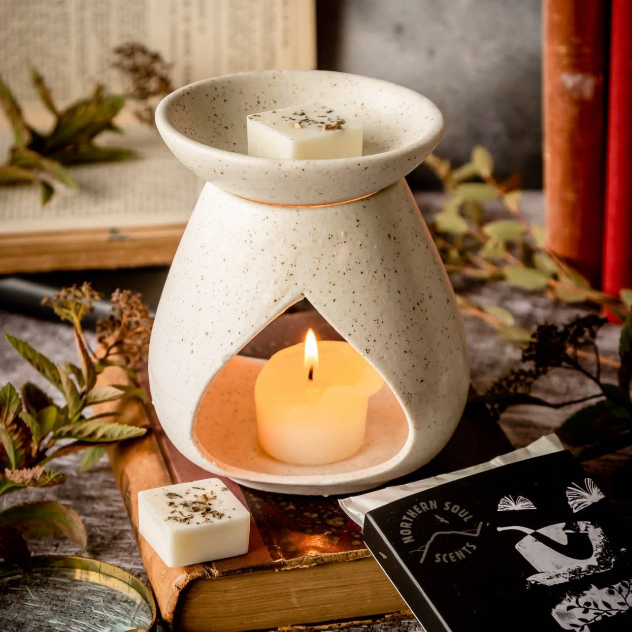 White ceramic oil burner with a lit candle on a rustic surface with books and leaves.