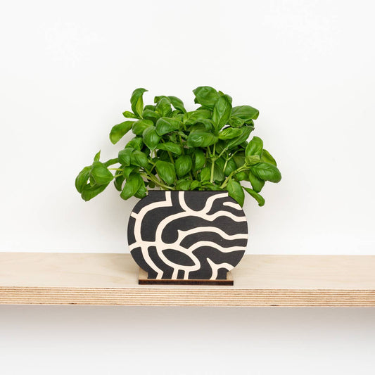 A black and white abstract patterned plant pot screen that hides ugly plant pots on a shelf with a plant behind it. 