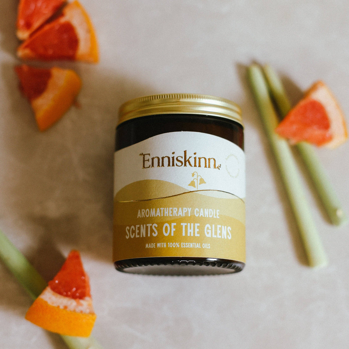 A jar of Enniskinn aromatherapy candle with the label 'Scents of the Glens' surrounded by slices of grapefruit and greenery on a light background.