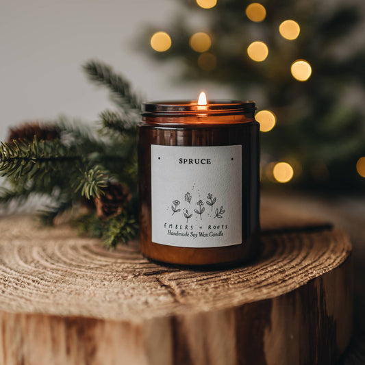 Candle in a jar labeled 'Spruce' on a wooden surface with blurred Christmas tree and lights in the background.