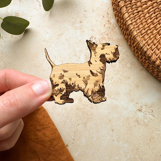A hand holding a wooden magnet in the shape of a Scottie Dog, with natural wood patterns.