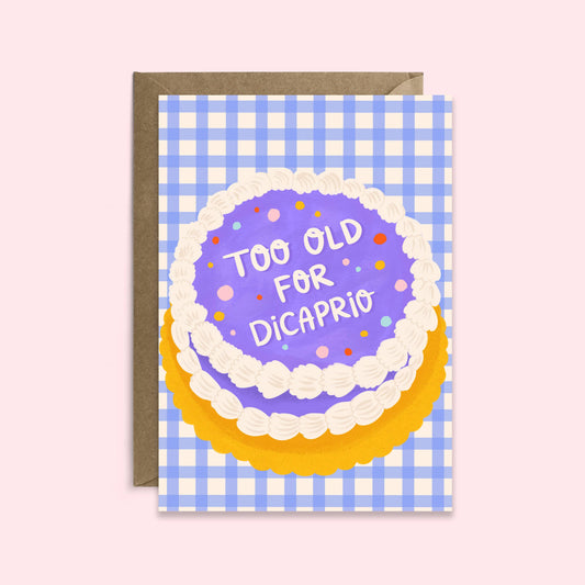 A humorous birthday card with a illustration of a cake and text 'TOO OLD FOR DICAPRIO' on it, presented on a blue checkered background with a brown kraft envelope.