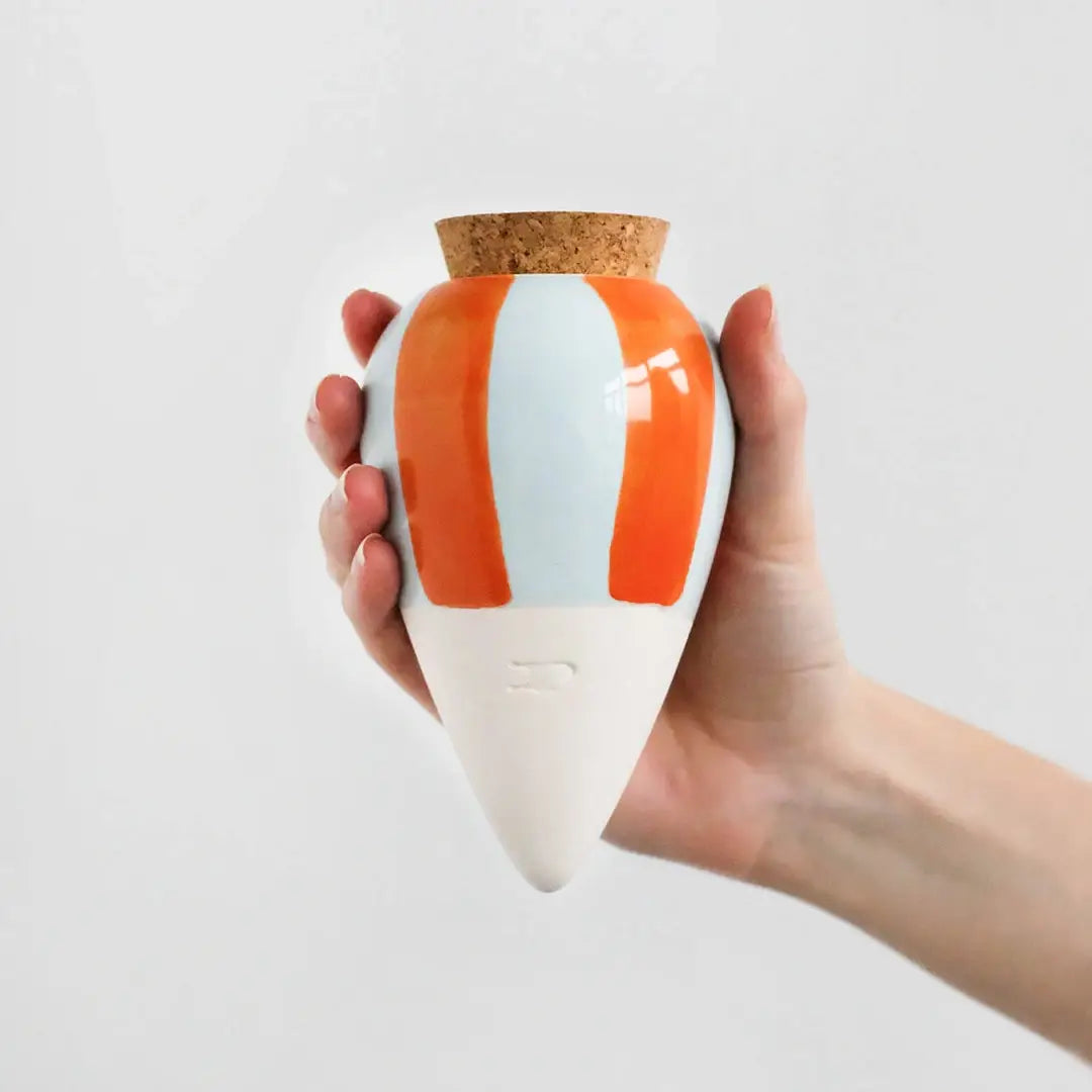 A hand holding a conical ceramic watering spike with orange and blue stripes, designed for indoor plants.