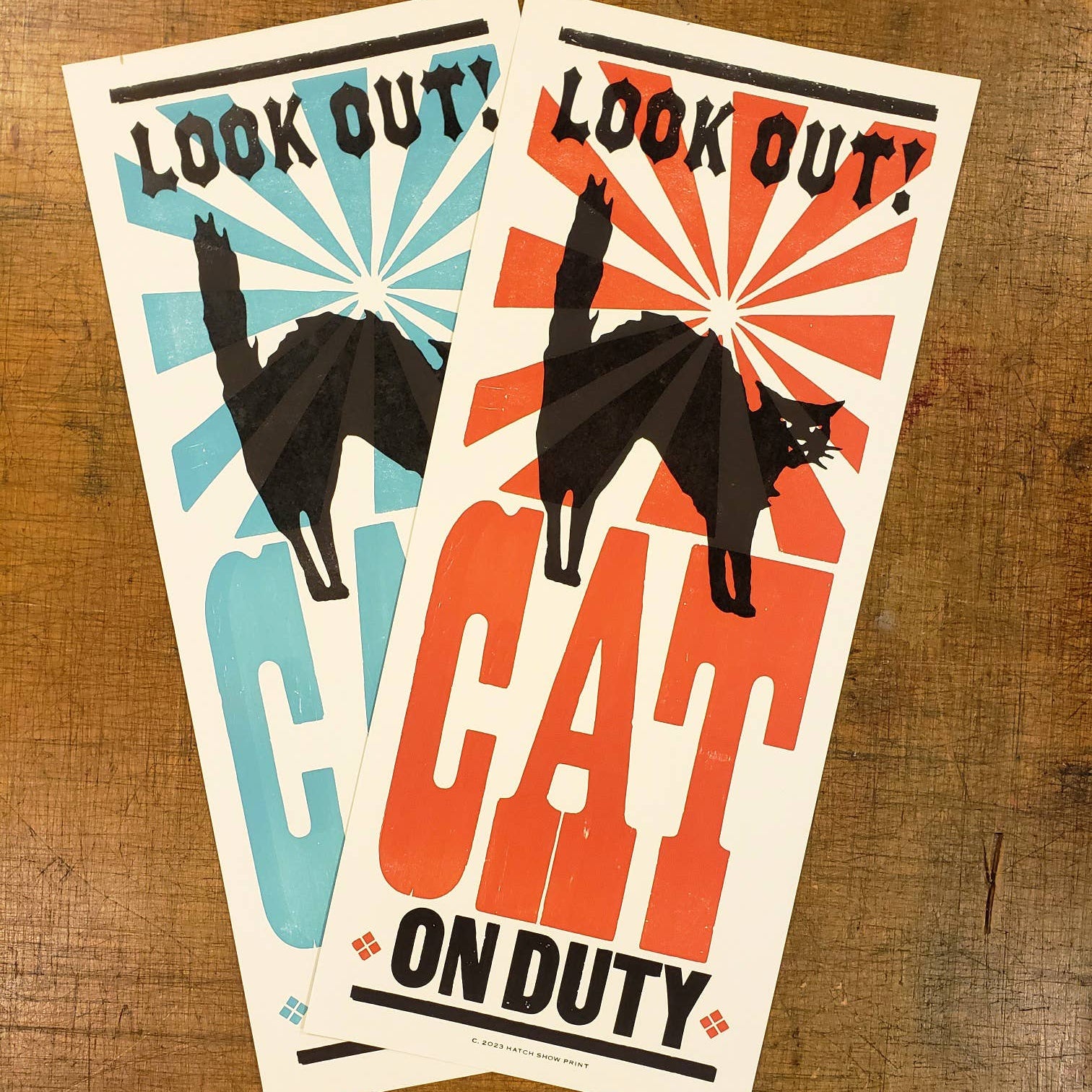 Two Hatch Show Print posters featuring a silhouette of a cat with the phrases 'Look out!' and 'Cat on duty' in bold lettering, one in blue and one in orange, on a wooden surface.
