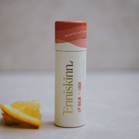 A stick of 'Enniskinn - Lugh' lip balm with orange and bergamot, placed upright beside a slice of orange, with the brand name and product scent visible on the packaging.