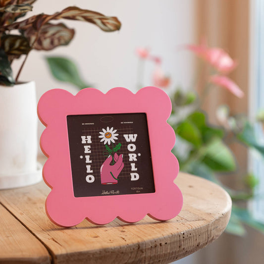 Pink scalloped photo frame with a black card inside on a wooden surface with plants in the background