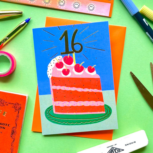 Greeting card with cake design and gold foil number 16, surrounded by stationery items on a green background