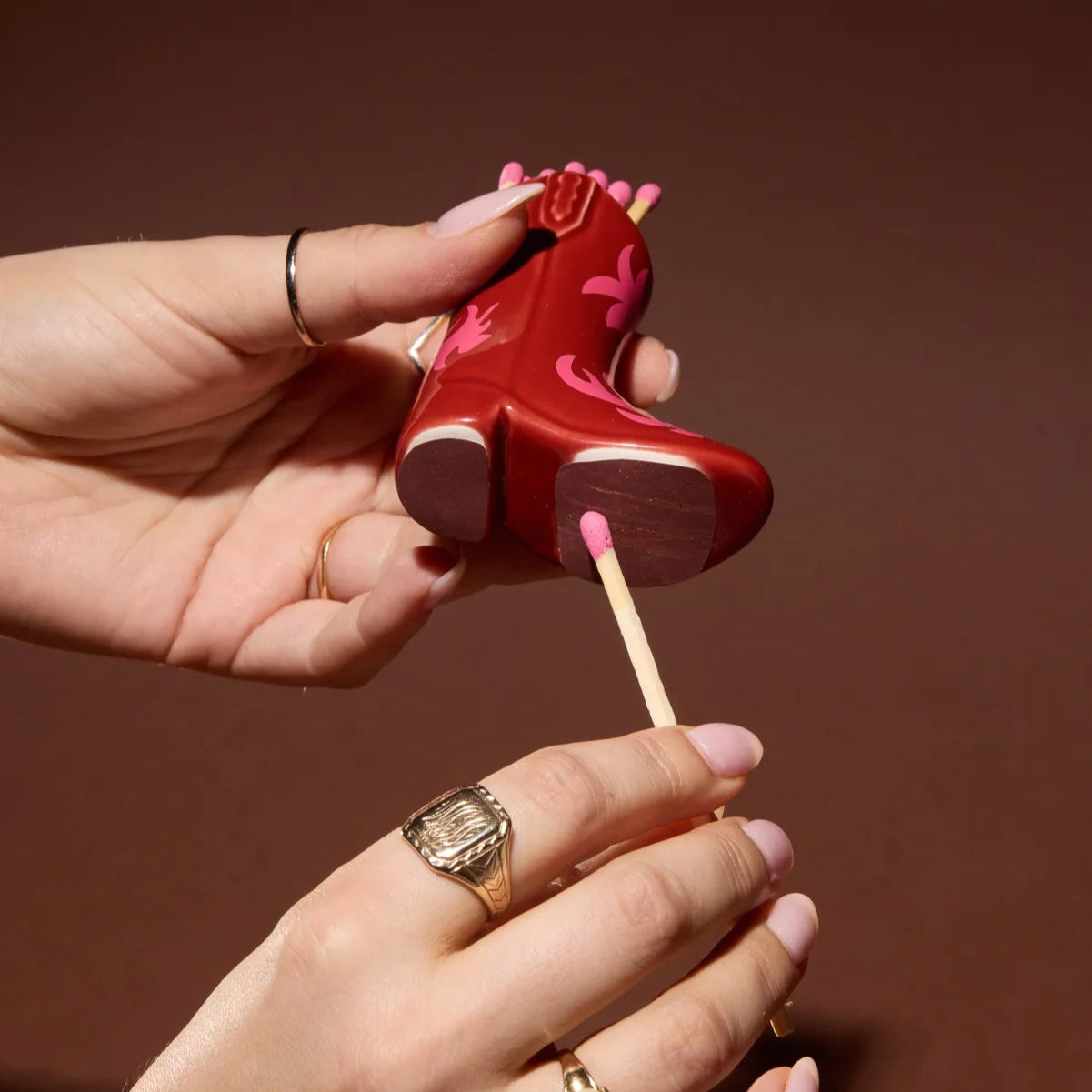 Retro style cowboy boot match holder in red and pink being held by hands striking a match against it
