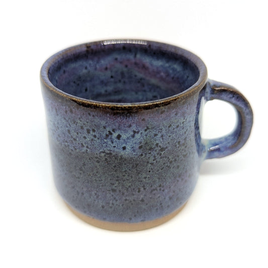 A handcrafted ceramic espresso mug with a purple and blue sunset color palette and a handle on the side, displayed against a neutral background.