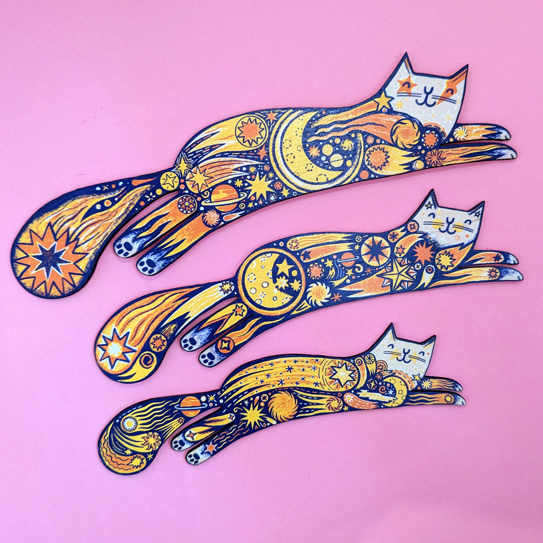 A set of three colorful wooden cat shapes with intricate patterns and designs, displayed against a pink background.