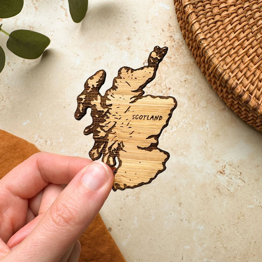 A hand holding a wooden magnet in the shape of a map of Scotland, with natural wood color and texture.