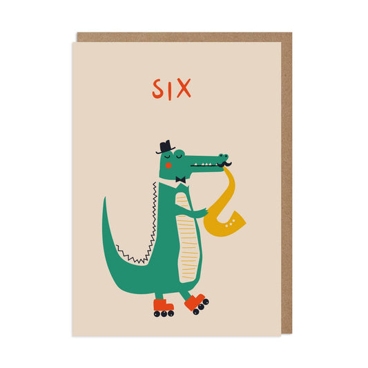 Greeting card featuring an illustrated crocodile wearing a hat and rollerblates, playing a saxophone. The card has a light background and includes a brown kraft envelope.