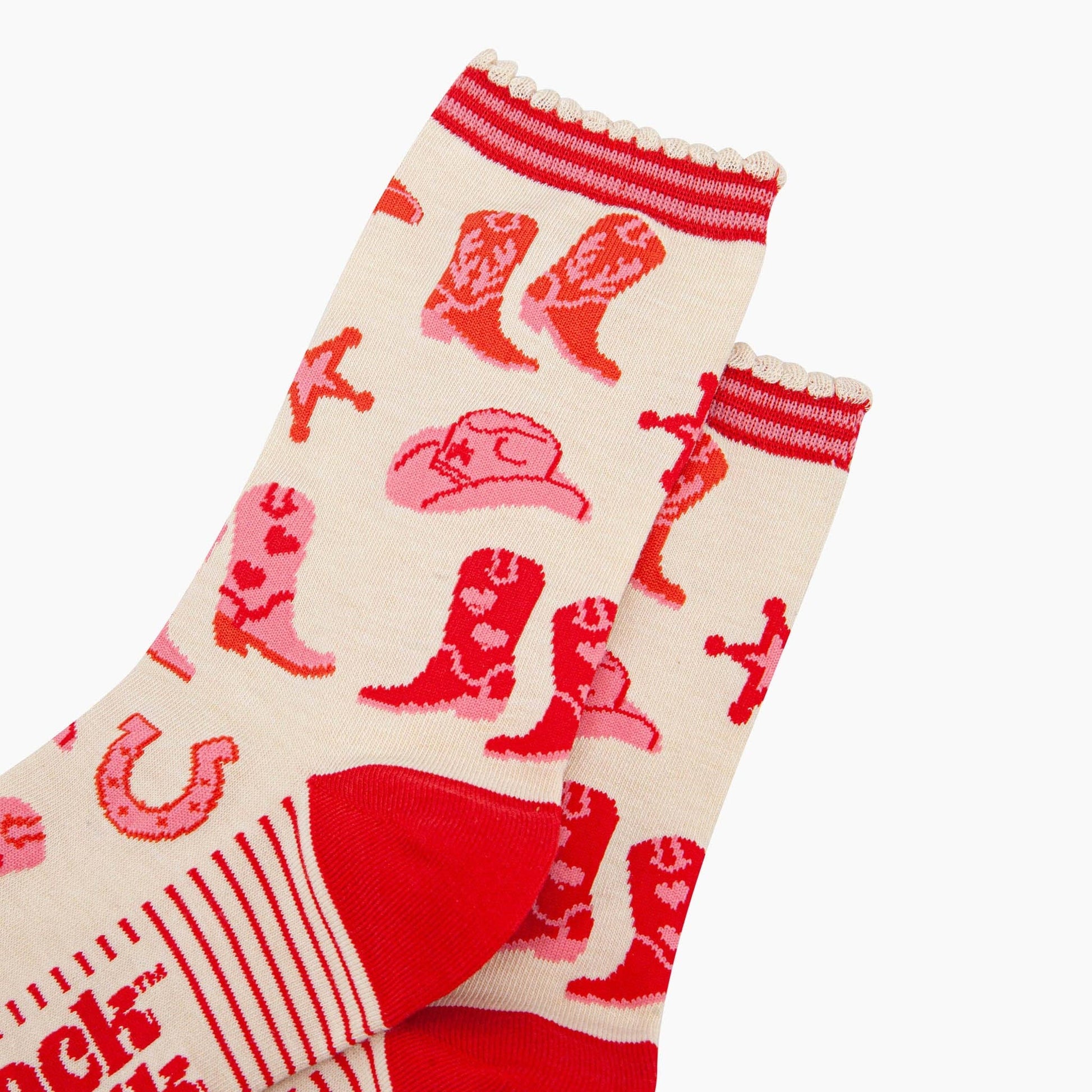 A pair of ankle socks with a western cowgirl theme, featuring red boots and accessories on a cream background with red stripes at the heel. Close up of ribbed cuff detail.