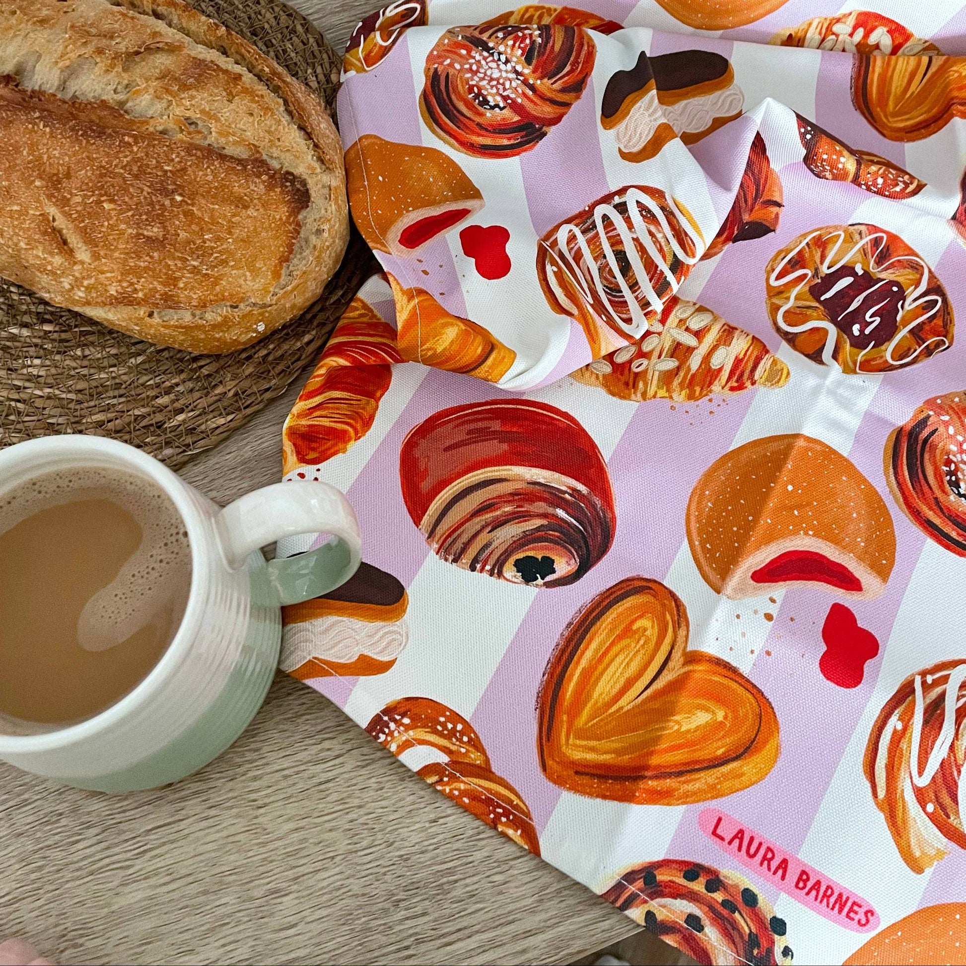 Tea towel with pastry design, bread, and mugs on a wooden surface