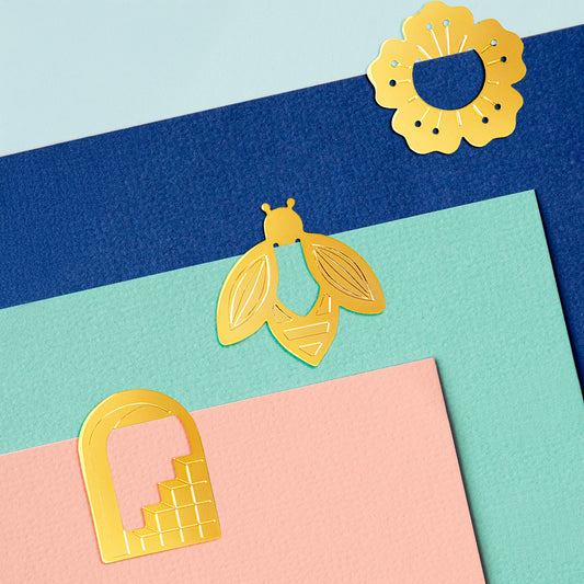 brass coloured paper clips in the shape of a doorway, a bee and a flower, clipped on to bright coloured card.