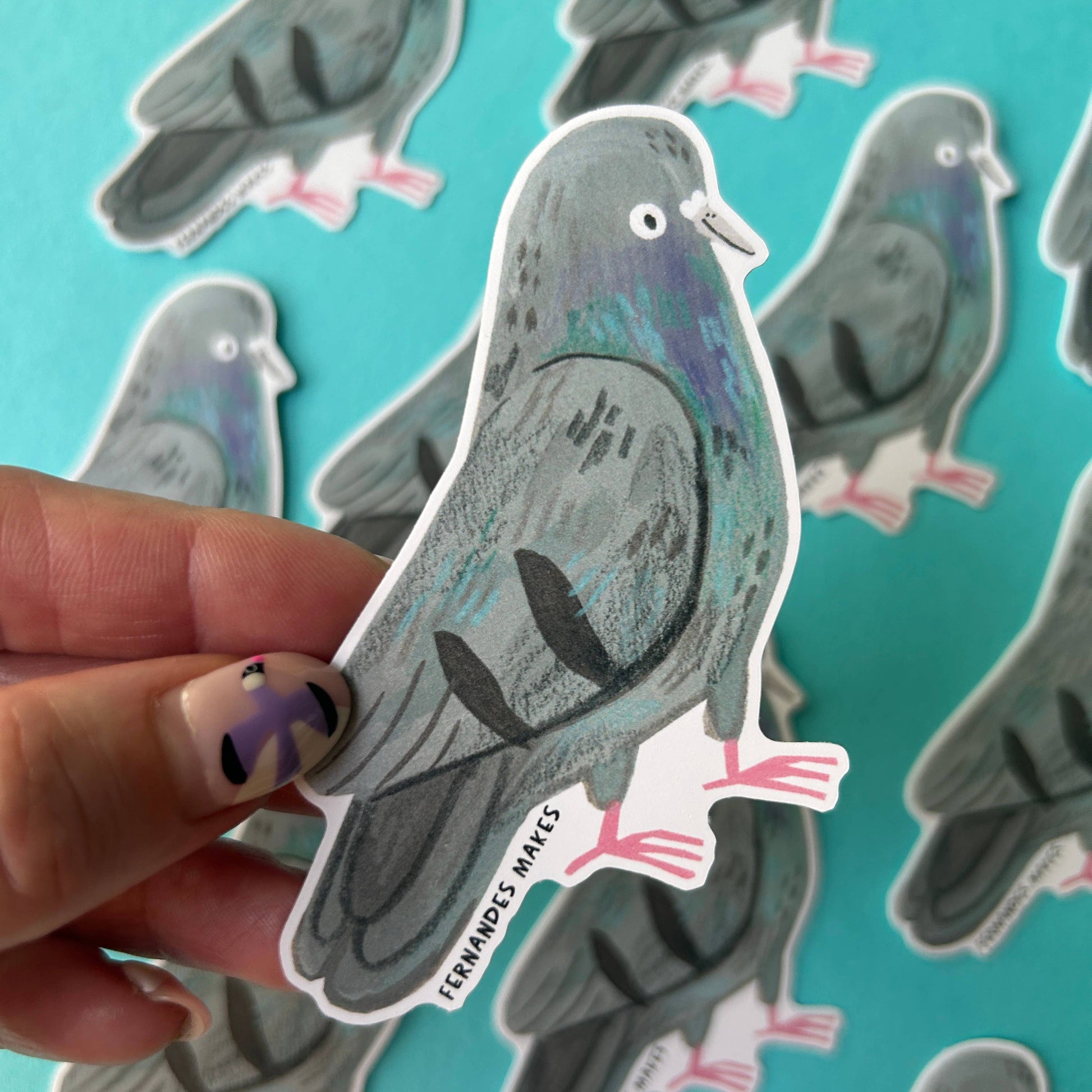 A hand holds a die-cut sticker shaped like a pigeon, with several matching pigeon stickers scattered on a bright turquoise background. 