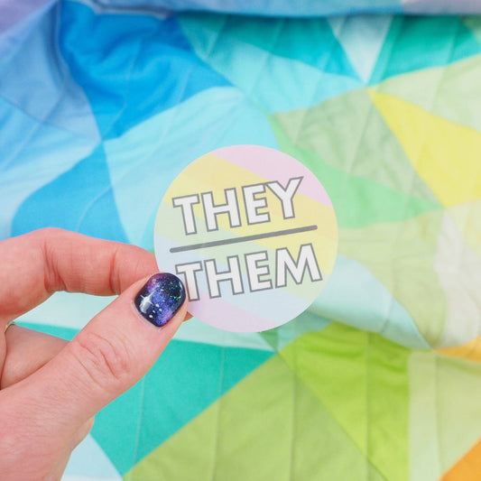 A hand holding a round pastel-colored vinyl sticker with the text 'THEY THEM' printed in the center, against a colorful geometric background.