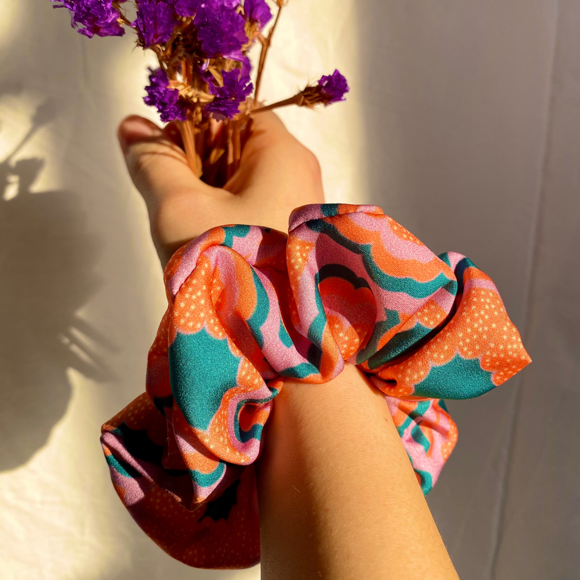 Hand holding a colorful scrunchie with purple flowers in the background