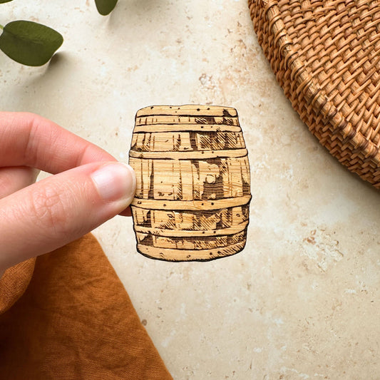 A hand holding a small, flat magnet with an illustration of a Scottish whisky barrel on it, placed on a light-colored surface with a woven object in the background.