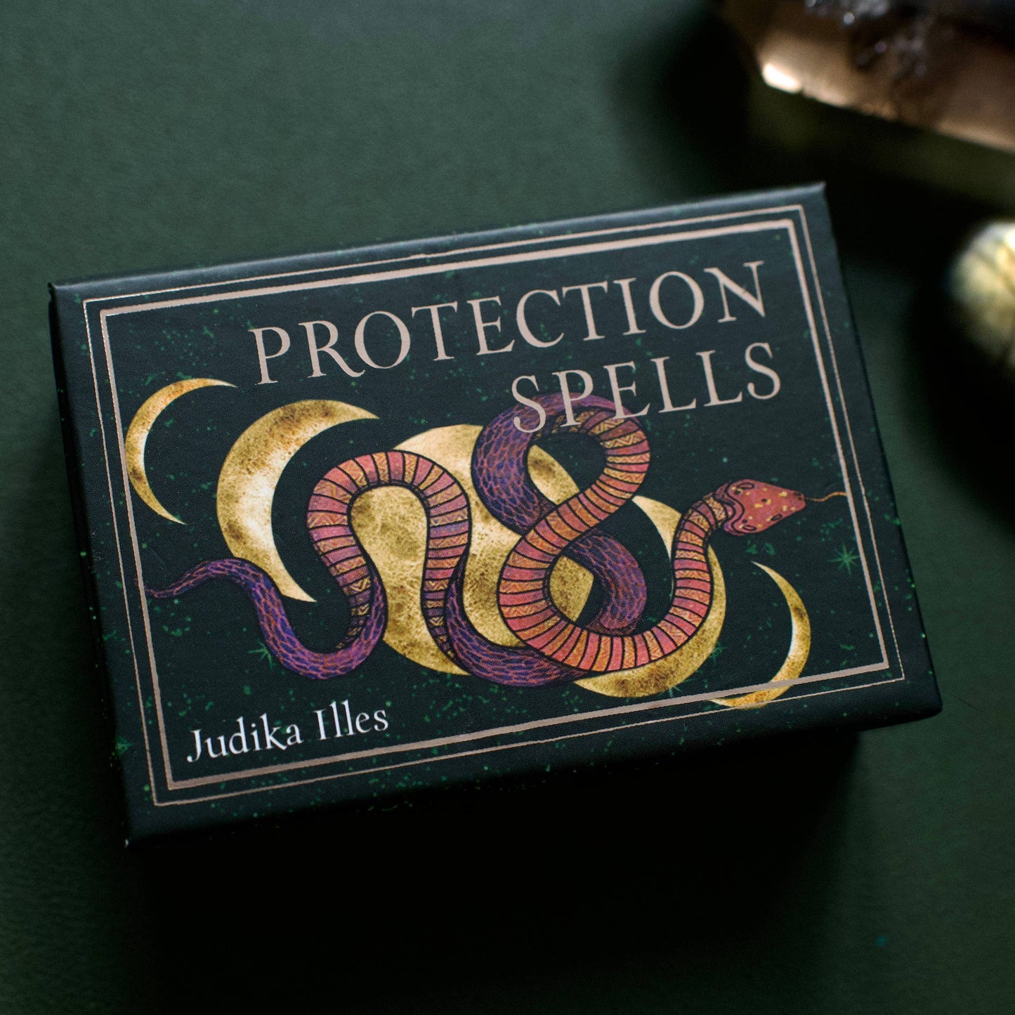 Box of 'Protection Spells' by Judika Illes on a dark green background with snake illustration on the packaging.