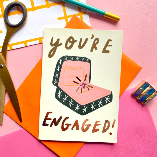 A greetings card with 'You're Engaged!' text and an engagement ring illustration, placed on a desk with scissors, a pen, and other stationery items.
