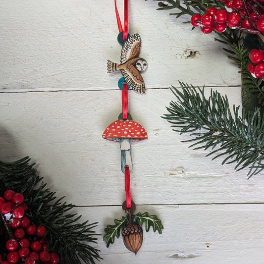 Decorative ornament with owl, mushroom, and acorn on a red ribbon against a white wooden background with greenery and berries.